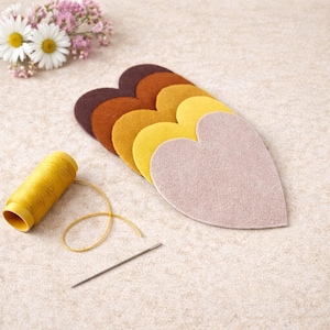 Puede incluir: Un kit de manualidades con piezas de fieltro en forma de corazón en tonos marrón, amarillo y rosa. También hay un carrete de hilo amarillo y una aguja, junto con un pequeño ramo de flores blancas y rosas. Materiales de manualidades.