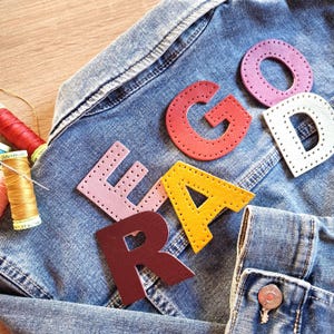May include: A blue denim jacket with colourful leather letters spelling "READ GOOD". The letters are red, pink, yellow, white, and burgundy. Sewing thread and spools are visible.