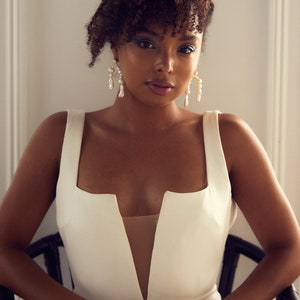 May include: A woman with curly brown hair wearing a white sleeveless dress with a deep V-neckline. She is wearing pearl earrings and has blue eyeliner.