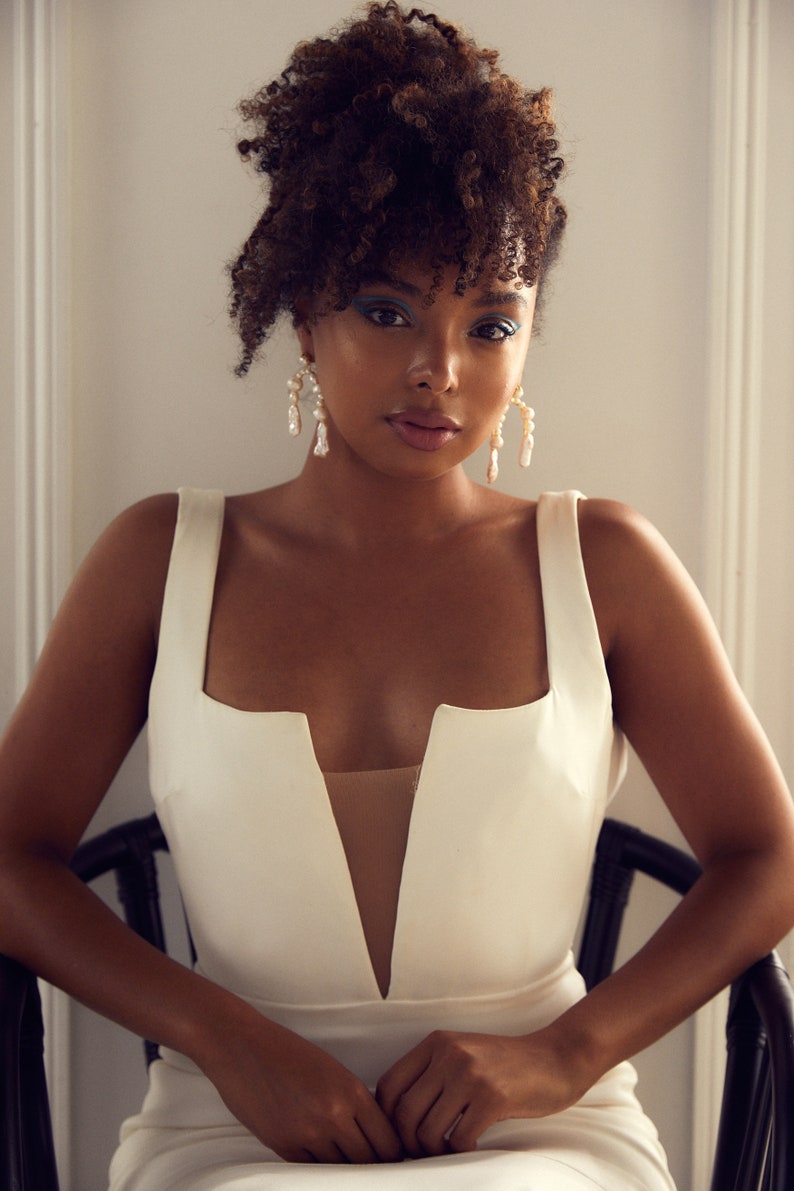 May include: A woman with curly brown hair wearing a white sleeveless dress with a deep V-neckline. She is wearing pearl earrings and has blue eyeliner.