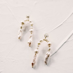 May include: A pair of gold hoop earrings with white pearl accents. The earrings are shaped like an arch and have a dangling pearl drop.
