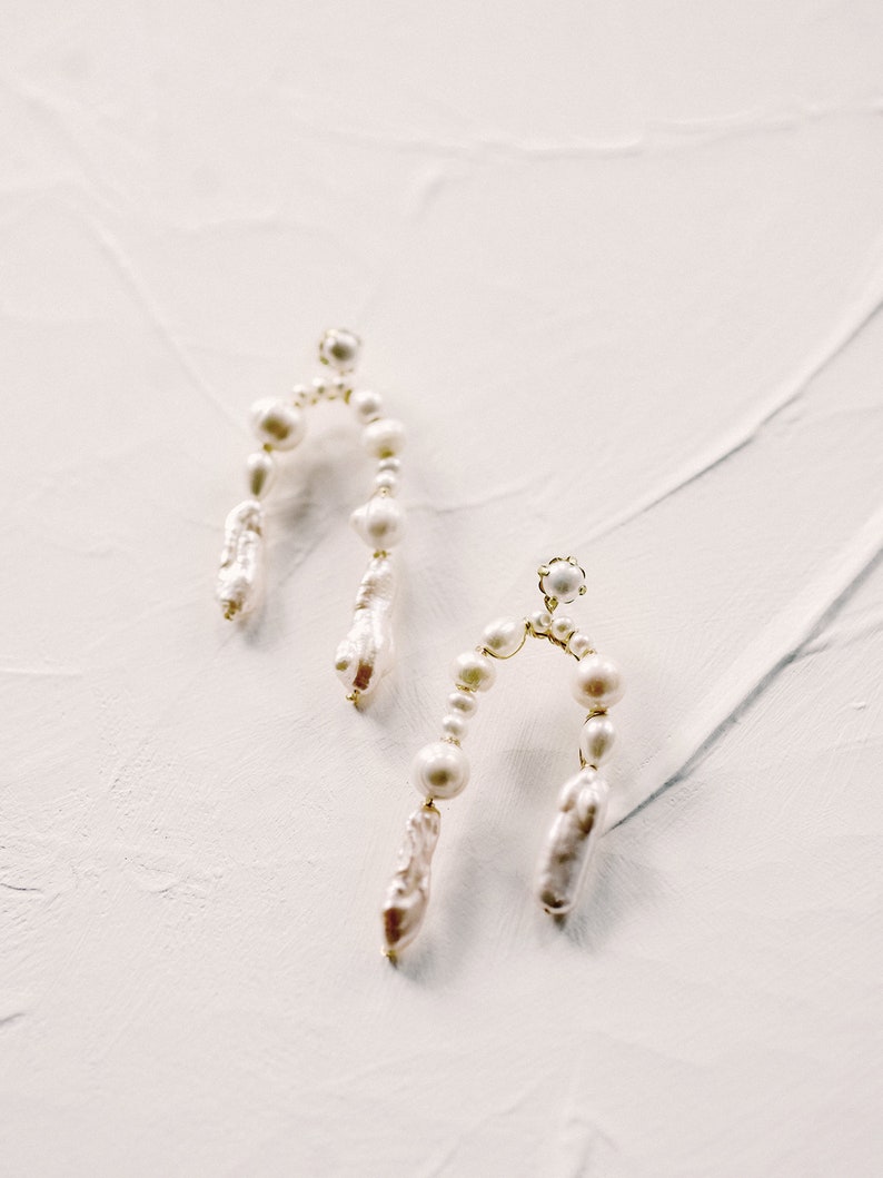 May include: A pair of gold hoop earrings with white pearl accents. The earrings are shaped like an arch and have a dangling pearl drop.