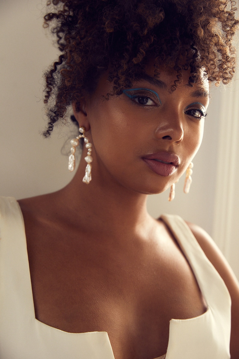 May include: A woman with curly brown hair wearing a white sleeveless top with a deep V-neckline. She is wearing large pearl and gold hoop earrings. Her makeup includes blue eyeliner.