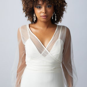 May include: A white wrap dress with sheer long sleeves and a V-neckline. The dress has a fitted bodice and a flowing skirt. The model is wearing pearl earrings.