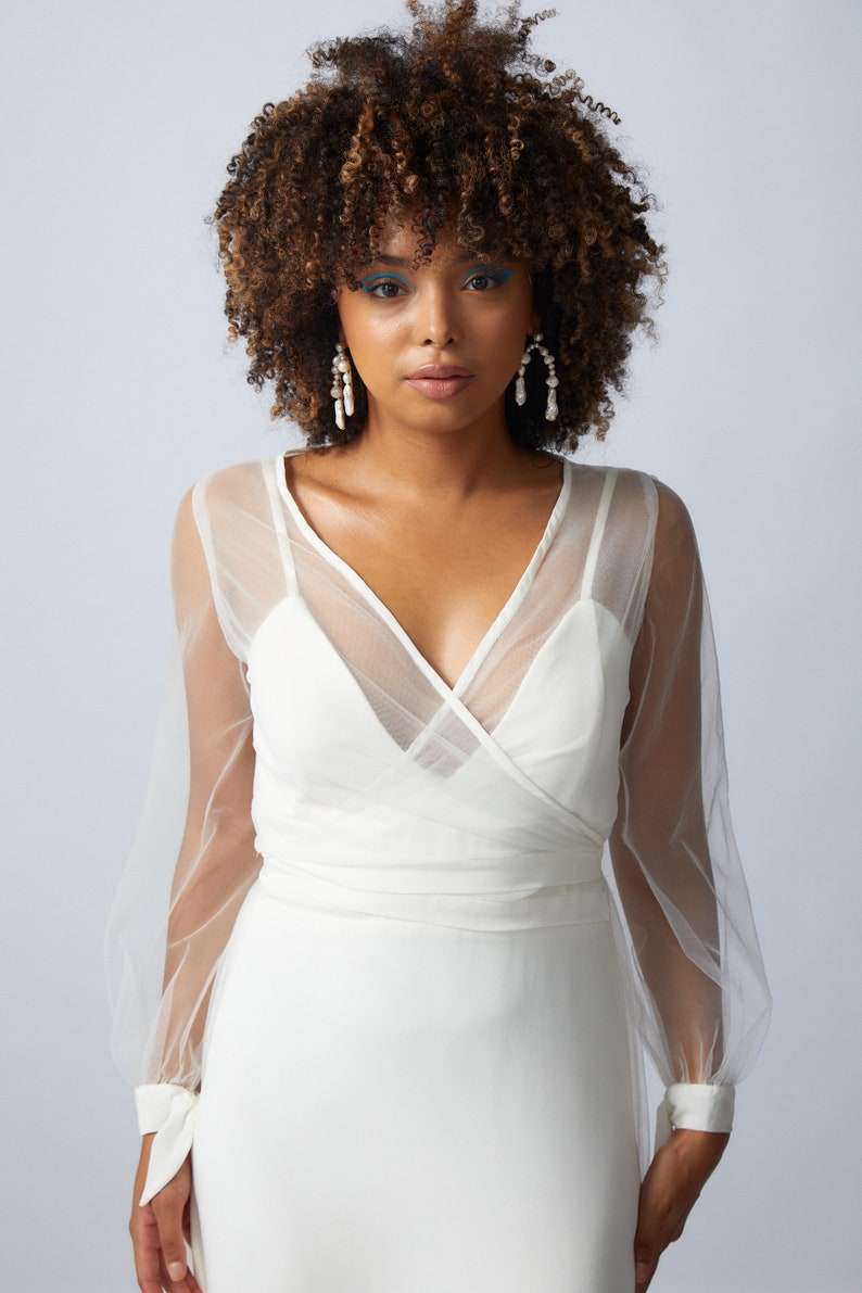 May include: A white wrap dress with sheer long sleeves and a V-neckline. The dress has a fitted bodice and a flowing skirt. The model is wearing pearl earrings.