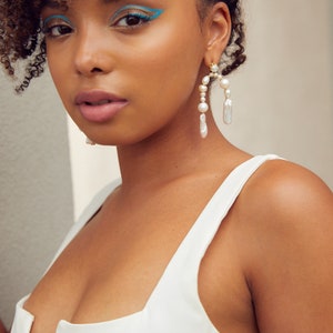 May include: A woman with curly hair wearing a white tank top and large pearl earrings. The earrings are gold and have a curved shape with multiple pearls.