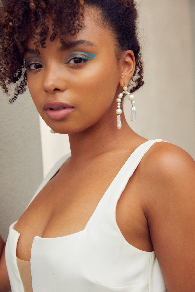 May include: A woman with curly hair wearing a white tank top and large pearl earrings. The earrings are gold and have a curved shape with multiple pearls.