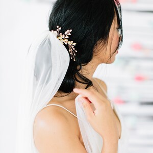 May include: A woman with black hair wearing a white veil and a gold hair accessory. The hair accessory is a floral design with small white accents. The veil is sheer and delicate, and the woman is wearing a white dress.