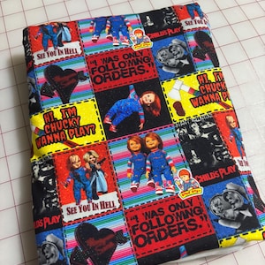 May include: A fabric item featuring a patchwork design with images of the Chucky doll from the Child's Play horror film series. The design includes text such as "See You In Hell" and "I Was Only Following Orders." The background has a striped pattern.