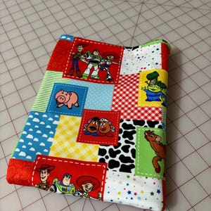 May include: A colorful, patchwork fabric item featuring various Toy Story characters. The design includes squares with images of Woody, Buzz Lightyear, and other characters, set against a background of red, blue, and white patterns.