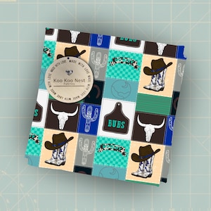 May include: A folded fabric with a patchwork design featuring cowboy boots, hats, cacti, and steer skulls in a repeating pattern. The fabric includes a tag that says "Koo Koo Nest Fabrics" and "Made with Love". The word "BUBS" is also visible.