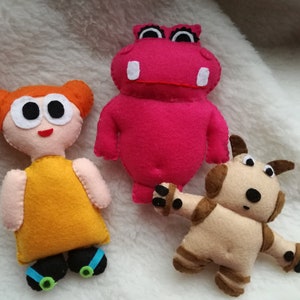 Puede incluir: Tres juguetes de fieltro hechos a mano: una niña con pelo naranja y un vestido amarillo, un hipopótamo rosa y una criatura parecida a un perro de color beige. Los juguetes están sobre una superficie blanca y esponjosa.