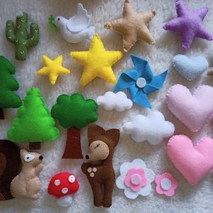 Puede incluir: Un conjunto de adornos de fieltro con una variedad de formas y diseños, incluyendo estrellas, corazones, árboles, una ardilla, un zorro, un hongo, un cactus, un pájaro, un molino de viento y nubes. Los adornos están hechos de fieltro y tienen una variedad de colores, incluyendo verde, marrón, rojo, rosa, azul, amarillo y blanco.
