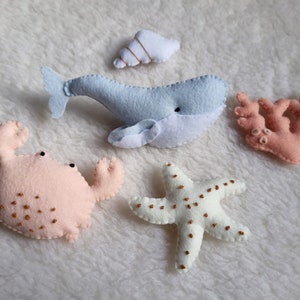 Puede incluir: Un conjunto de cinco juguetes de animales marinos de fieltro, que incluyen una ballena azul, un cangrejo rosa, una estrella de mar blanca, un coral rosa y una concha blanca.