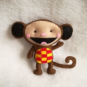 Può includere: Un giocattolo di scimmia di feltro marrone con orecchie nere, una camicia a quadri rossi e gialli e un grande sorriso.