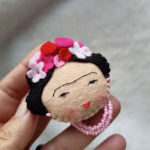 May include: A felt doll with black hair, a pink and white flower crown, and a pink pearl necklace. The doll has a brown face with a black unibrow and red lips.