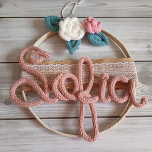 Può includere: Decorazione da parete fatta a mano con il nome "Sofia" in lettere rosa lavorate a maglia. Il design include un cerchio color crema, dettagli in iuta e pizzo e fiori all'uncinetto bianchi, rosa e turchese.