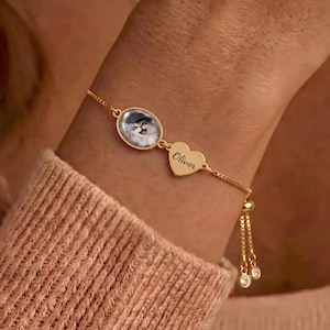 Könnte beinhalten: Ein goldfarbenes, verstellbares Kettenarmband mit einem ovalen Anhänger, der das Bild einer Katze mit Mütze zeigt, und einem herzförmigen Anhänger mit der Gravur Oliver. Das Armband hat zwei herabhängende Enden mit kleinen, klaren Steinen.
