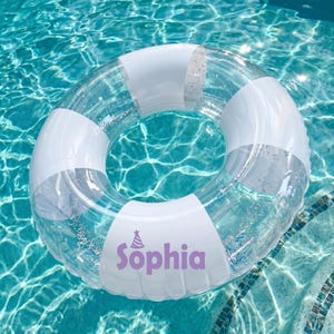 Puede incluir: Un flotador de piscina inflable circular y transparente con secciones blancas alternas y detalles de purpurina plateada en el interior. Una sección blanca tiene impreso un icono de gorro de fiesta morado sobre el nombre Sophia.