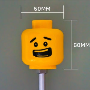 K&ouml;nnte beinhalten: Ein gelber LEGO Minifigurenkopf mit einem l&auml;chelnden Gesicht und hochgezogenen Augenbrauen. Der Kopf ist 5 cm breit und 6 cm hoch.