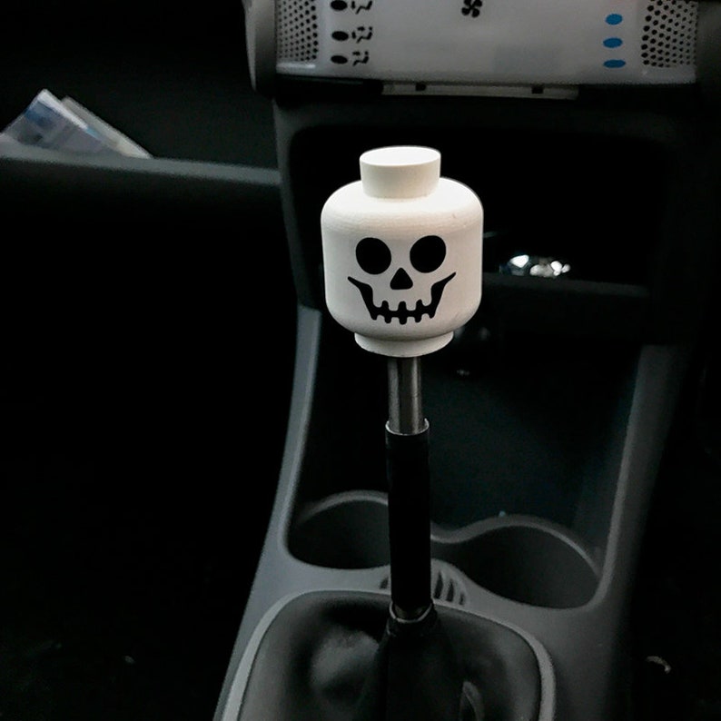 May include: A white plastic gear shift knob with a black skull design. The knob is attached to a black gear shift lever.