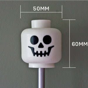 May include: White Lego minifigure head with a black skull face. The head is 2 inches wide and 2.3 inches tall.