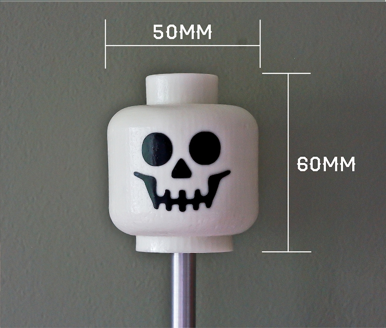 May include: White Lego minifigure head with a black skull face. The head is 2 inches wide and 2.3 inches tall.
