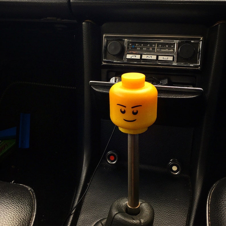 K&ouml;nnte beinhalten: Ein gelber Lego-Figurenkopf als Schaltknauf f&uuml;r ein Auto. Die Figur hat einen ernsten Ausdruck mit zusammengezogenen Augenbrauen und einem kleinen L&auml;cheln.