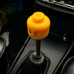 K&ouml;nnte beinhalten: Ein gelber Lego-Figurenkopf-Schalthebelknauf f&uuml;r ein Auto. Die Figur hat einen ernsten Ausdruck mit zusammengezogenen Augenbrauen.