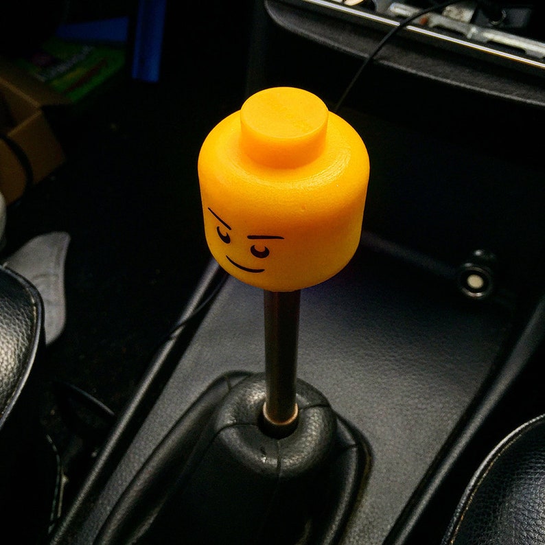 K&ouml;nnte beinhalten: Ein gelber Lego-Figurenkopf-Schalthebelknauf f&uuml;r ein Auto. Die Figur hat einen ernsten Ausdruck mit zusammengezogenen Augenbrauen.