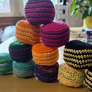 May include: A collection of ten round, crocheted hacky sacks stacked in a pyramid formation. The items feature various color combinations including solid purple, orange, black and yellow stripes, and multi-colored speckled yarn patterns.