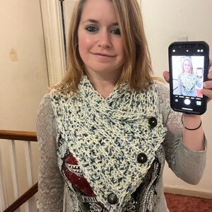 May include: A crocheted cowl scarf in cream and blue, fastened with three large brown buttons. The scarf has a textured, open weave design. The person in the image is holding a phone showing a picture of the scarf.