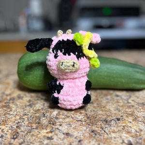 May include: A handmade crochet cow figurine in pink, black, and beige. The cow has black spots, a beige snout, and small yellow and green accents. The cow is approximately 4 inches tall.