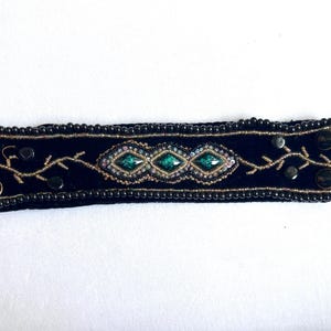 May include: A black velvet cuff bracelet featuring three central emerald green diamond-shaped stones surrounded by intricate gold embroidery and dark beads. The edges are lined with small black beads, and the ends include two black fabric loops for closure.