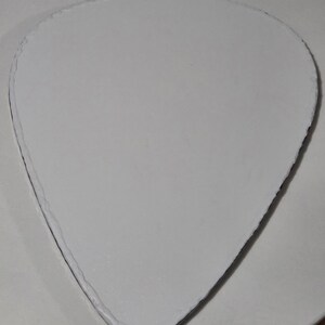 Puede incluir: Un objeto blanco en forma de p&uacute;a de guitarra, con una superficie texturizada y un borde oscuro. El objeto est&aacute; centrado en una superficie circular blanca. La forma de la p&uacute;a es un dise&ntilde;o com&uacute;n para accesorios de guitarra.