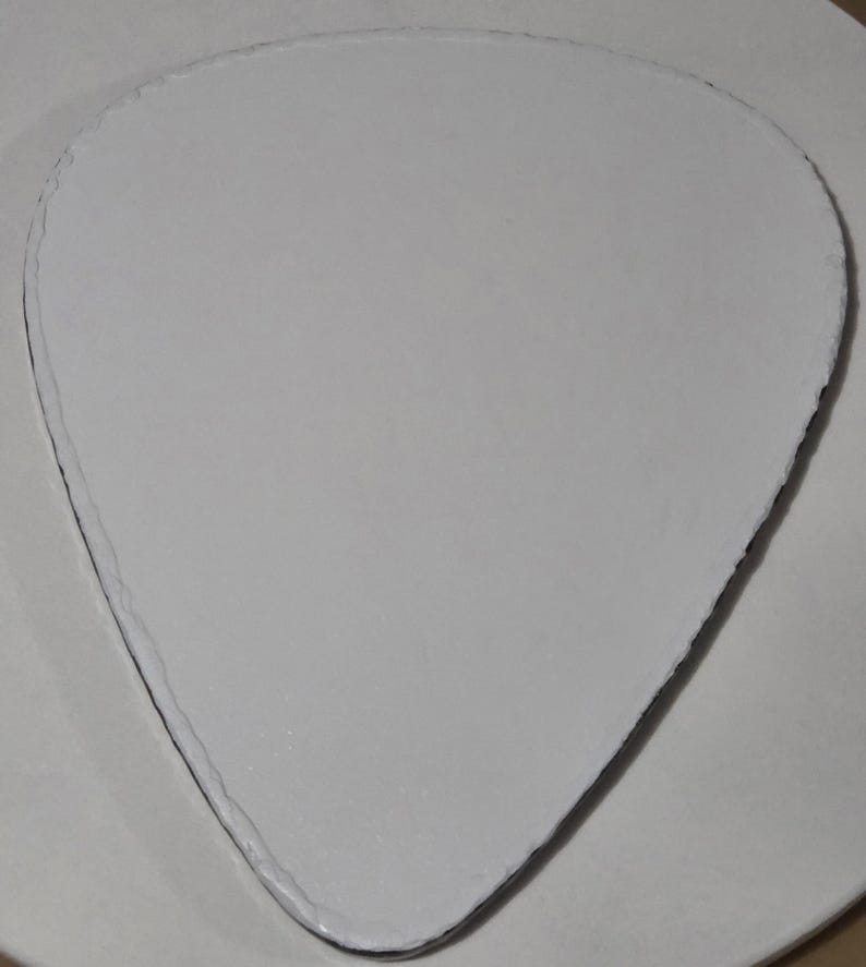 Puede incluir: Un objeto blanco en forma de p&uacute;a de guitarra, con una superficie texturizada y un borde oscuro. El objeto est&aacute; centrado en una superficie circular blanca. La forma de la p&uacute;a es un dise&ntilde;o com&uacute;n para accesorios de guitarra.