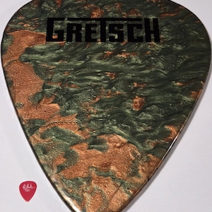 Puede incluir: Una p&uacute;a de guitarra grande y personalizada con un dise&ntilde;o en espiral verde y cobrizo. La palabra "GRETSCH" est&aacute; impresa en negro en la parte superior. Una peque&ntilde;a p&uacute;a de guitarra roja est&aacute; en la esquina inferior izquierda.