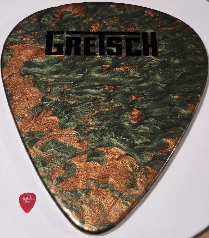 Puede incluir: Una p&uacute;a de guitarra grande y personalizada con un dise&ntilde;o en espiral verde y cobrizo. La palabra "GRETSCH" est&aacute; impresa en negro en la parte superior. Una peque&ntilde;a p&uacute;a de guitarra roja est&aacute; en la esquina inferior izquierda.