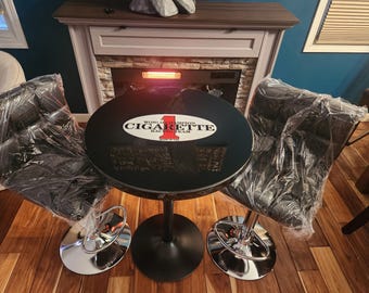 May include: A black table with a round black top featuring a white oval sticker with the text "World 1 Champion Cigarette Racing Team". The table has a black metal base and is surrounded by two black bar stools with chrome bases.
