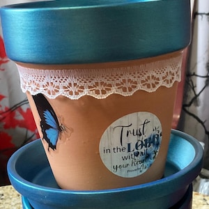 May include: A terracotta flower pot with a blue rim and matching saucer. The pot is decorated with a white lace trim, a blue butterfly sticker, and a round sticker with the text "Trust in the Lord with all your heart."