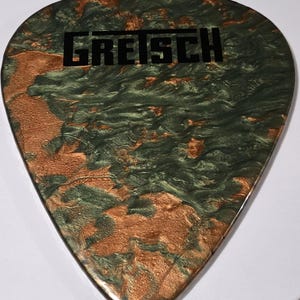 Puede incluir: Una p&uacute;a de guitarra con un dise&ntilde;o veteado en tonos verdes y cobrizos. La palabra "GRETSCH" est&aacute; impresa en negro en el centro. La p&uacute;a tiene un acabado liso y brillante.