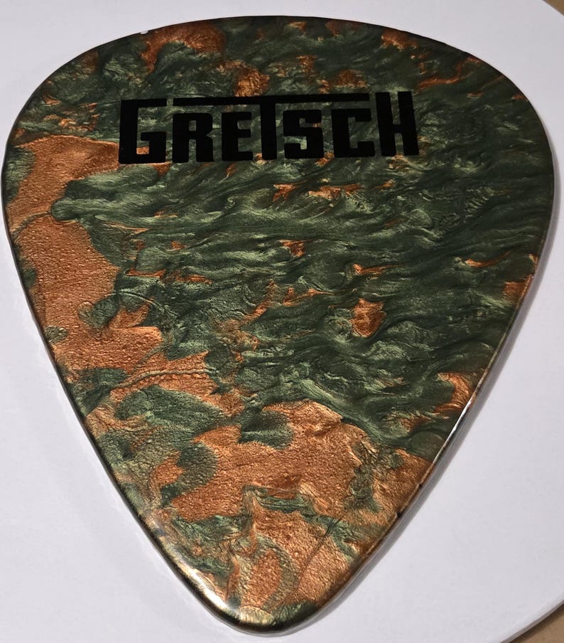 Puede incluir: Una p&uacute;a de guitarra con un dise&ntilde;o veteado en tonos verdes y cobrizos. La palabra "GRETSCH" est&aacute; impresa en negro en el centro. La p&uacute;a tiene un acabado liso y brillante.