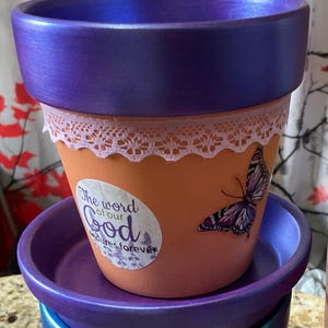 May include: A decorative terracotta flower pot with a purple rim and saucer. The pot features a white lace trim, a butterfly decal, and a sticker with the text "The word of our God endures forever."