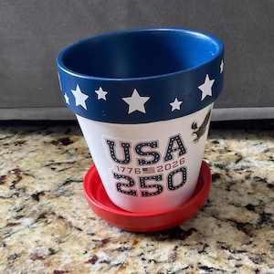 May include: A patriotic flower pot with a blue rim and white stars. The pot features the text "USA 250" with the years 1776 and 2026, and an eagle graphic. It sits on a red saucer.