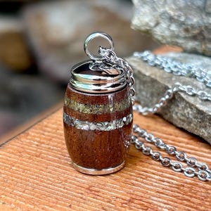 May include: A barrel-shaped pendant necklace with a silver chain. The pendant is made of dark brown wood with bands of lighter-colored stone and a silver cap. The necklace is displayed on a wooden surface with a blurred stone background.