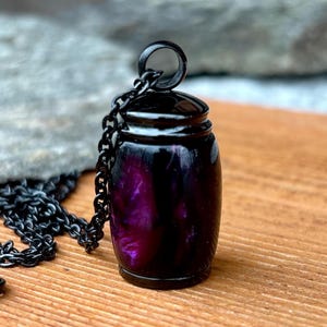 May include: A black jar-shaped pendant necklace with a chain. The jar has a dark purple and black marbled design. The necklace is set on a wooden surface with a blurred stone background.