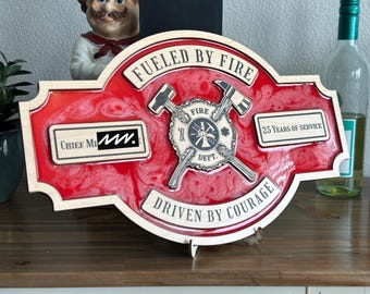 May include: A red and white wooden plaque with the text "Fueled by Fire" and "Driven by Courage". The plaque also features a firefighter's helmet and crossed axes, and the text "Chief M.M." and "25 Years of Service".