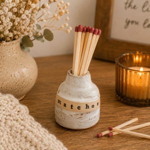 May include: A small, speckled white ceramic vessel with a narrow neck and a beige band featuring the black text "matches" printed on it. The vessel is filled with wooden matches that have dark red tips, with two additional matches lying nearby.