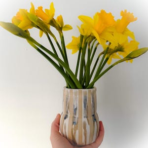 May include: A hand holds a ceramic vase filled with yellow daffodils. The vase has beige and blue vertical stripes. The daffodils have green stems and are in full bloom, set against a white backdrop.