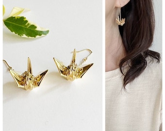 May include: Gold-colored origami crane earrings. The earrings feature a detailed crane design, with the wings and head clearly defined. The earrings are shown on a white background, with a close-up of the earrings and a person wearing one.